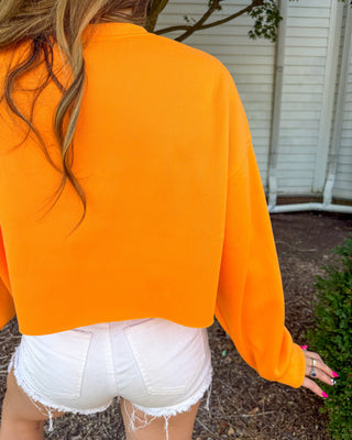 A person viewed from behind wearing a bright neon orange long-sleeve cropped sweatshirt and white denim shorts in an outdoor setting.