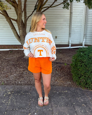 A woman stands outdoors on a paved walkway in front of a white house, wearing a white long-sleeve sweatshirt with orange 'Tennessee Volunteers' text and graphics, paired with bright orange shorts and tan sandals.