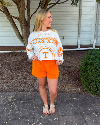 A woman stands outdoors on a paved walkway in front of a white house, wearing a white long-sleeve sweatshirt with orange 'Tennessee Volunteers' text and graphics, paired with bright orange shorts and tan sandals.