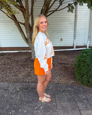 A woman stands outdoors on a paved walkway in front of a house, wearing a white cropped sweatshirt with orange lettering and bright orange shorts.