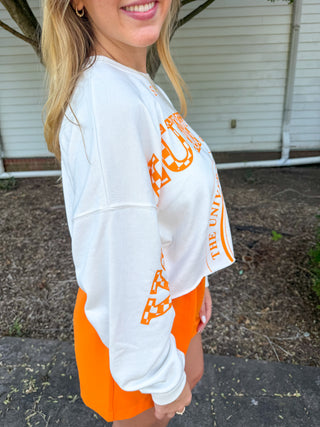 A woman wearing a white cropped sweatshirt with orange collegiate-style lettering and bright orange shorts, standing outdoors in front of a house wall.