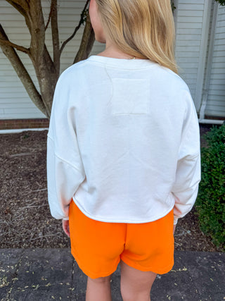 A person standing outdoors in front of a house, shown from the back wearing a white cropped long-sleeve sweatshirt and bright orange shorts.