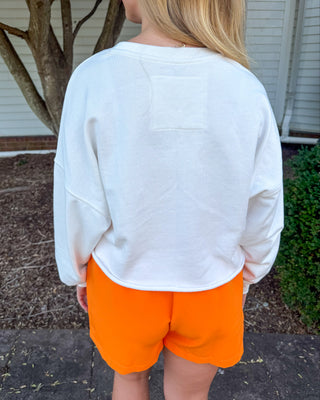 A person standing outdoors in front of a house, shown from the back wearing a white cropped long-sleeve sweatshirt and bright orange shorts.
