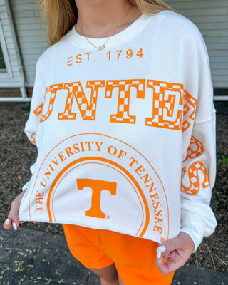 A woman wearing a white crewneck sweatshirt with orange University of Tennessee graphics and bright orange shorts, standing outdoors in front of a house.