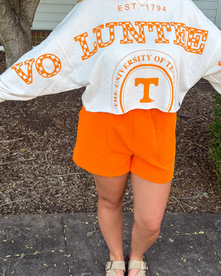 A woman stands outdoors on a stone walkway wearing a white long-sleeve sweatshirt with orange lettering that reads 'VO LUNTECH' and 'Tennessee Volunteers,' paired with bright orange shorts.