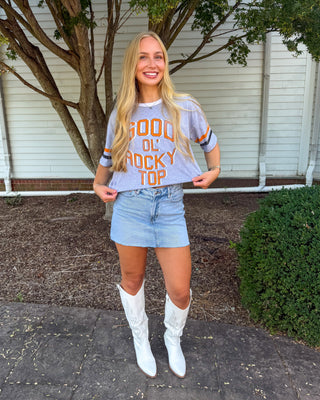 A woman stands outdoors in front of a house and tree, wearing a light gray graphic T-shirt with orange text reading 'GOOD OR ROCKY TOP', a light blue denim mini skirt, and white cowboy boots.