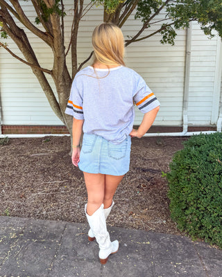A woman is shown from behind wearing a light blue heathered t-shirt with orange and navy sleeve stripes, a light blue denim mini skirt, and white cowboy boots, standing outdoors in front of a white house and tree.