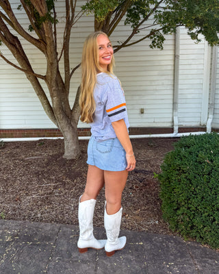 A woman stands outdoors on a paved walkway in front of a white house, wearing a light blue short-sleeve shirt with orange and navy stripes on the sleeve, a light blue denim mini skirt, and white cowboy boots.