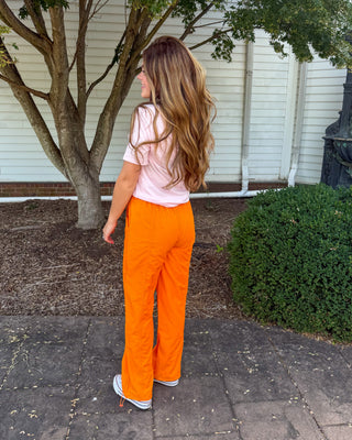 A woman stands outdoors on a stone walkway in front of a white house, wearing bright orange wide-leg pants, a light pink short-sleeve top, and white sneakers.