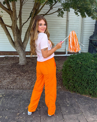 A woman stands outdoors on a paved walkway in front of a house, wearing bright orange pants and a light pink t-shirt, holding a pink and orange striped mop handle.