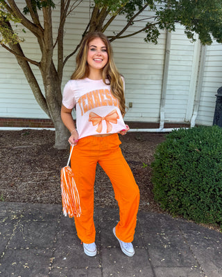 A woman stands outdoors on a paved walkway in front of a white building and tree, wearing a white graphic T-shirt with orange lettering and bow details, bright orange pants, and white sneakers. She is holding a white and orange pom-pom.