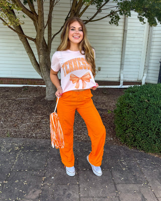 A woman stands outdoors on a paved walkway in front of a white building and tree, wearing a white graphic T-shirt with orange lettering and bow details, bright orange pants, and white sneakers. She is holding a white and orange pom-pom.