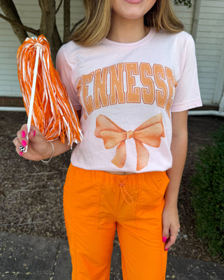 A woman wearing a light pink graphic t-shirt with 'TENNESSEE' and a bow design in orange, paired with bright orange drawstring pants, holding an orange and white cheer pom-pom outdoors.