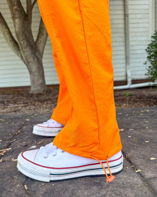 Bright orange pants with drawstring hems worn with white low-top sneakers on a paved outdoor walkway.