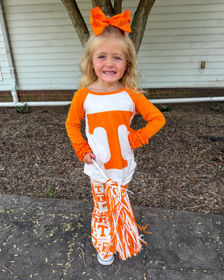 A young girl stands outdoors on a paved walkway in front of a house, wearing a Tennessee Volunteers cheerleader outfit. The outfit consists of a long-sleeve raglan shirt with orange and white sections and a large white 'T' on the front, matching orange and white pom-poms, and an orange hair bow.