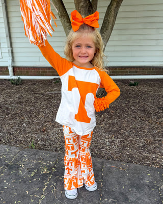 A young girl stands outdoors on a paved walkway in front of a house, wearing an orange and white Tennessee Volunteers cheerleader outfit with a large orange bow in her hair and holding an orange pom-pom.