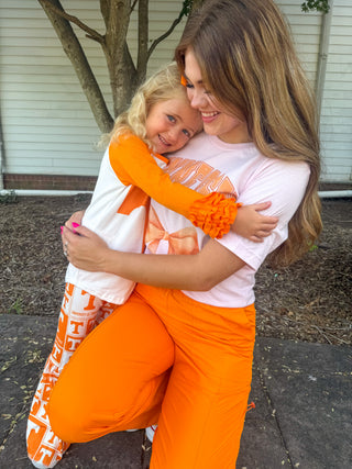 A woman and a young girl hugging outdoors. Both are wearing matching white t-shirts with orange collegiate-style lettering and bright orange pants.
