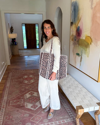A woman stands in a home hallway wearing a white long-sleeve top and wide-leg pants, carrying a large leopard-print tote bag with white trim. The setting features a patterned rug, a woven bench, and a large abstract painting on the wall.