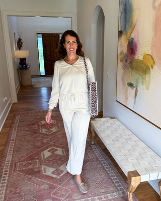 A woman wearing a cream-colored two-piece lounge set with a drawstring waist and wide-leg pants, walking in a hallway with a patterned rug, a woven bench, and a large abstract painting.