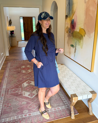 A woman wearing a navy blue shift dress, beige flat shoes, and a baseball cap stands in a home hallway next to a bench and a large abstract painting.