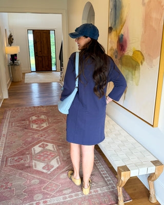 A woman stands in a hallway wearing a navy blue long-sleeve dress, a navy baseball cap, and a light blue shoulder bag, viewed from behind. The setting features a patterned rug, a bench, and a large abstract painting on the wall.
