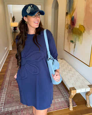 A woman wearing a navy blue long-sleeve dress with a large bow detail on the front, a navy baseball cap, and a light blue shoulder bag, standing in a hallway.