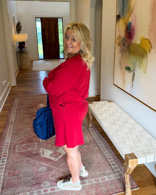 A woman wearing a bright red oversized short-sleeve dress stands in a home hallway, holding a blue bag. The hallway features a large abstract painting on the wall, a white bench, and a patterned rug.
