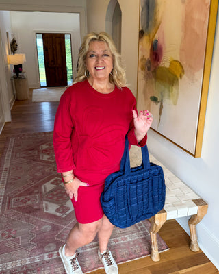 A woman stands in a home hallway wearing a matching red long-sleeve top and shorts set, holding a blue quilted tote bag. The background features a patterned rug, wooden floors, and a large abstract painting on the wall.