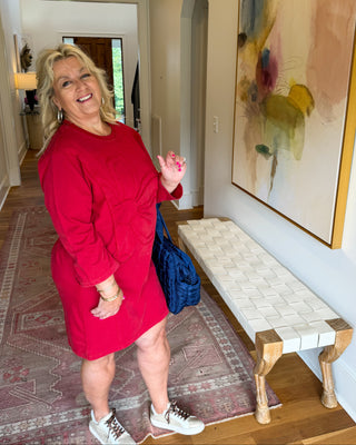 A woman wearing a bright red long-sleeve dress stands in a home hallway next to a woven bench and a large abstract painting.