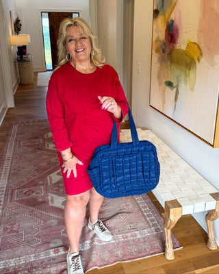 A woman stands in a home hallway wearing a red long-sleeve dress and holding a blue quilted shoulder bag. The background features a patterned rug, a bench, and a large abstract painting on the wall.