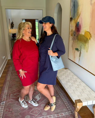 Two women posing together in a home hallway. The woman on the left wears a bright red long-sleeve dress and white sneakers, while the woman on the right wears a navy blue long-sleeve dress, a navy baseball cap, and a light blue shoulder bag.