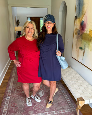 Two women posing together in an indoor hallway setting. The woman on the left wears a bright red dress with white sneakers, and the woman on the right wears a navy blue dress with a light blue shoulder bag and a dark cap.