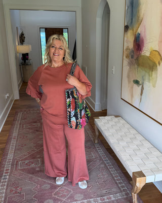 A woman stands in a home hallway wearing a matching dusty rose pink lounge set consisting of a short-sleeve top and wide-leg pants, holding a colorful patterned shoulder bag.