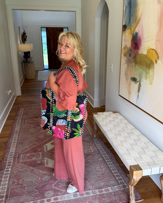 A woman stands in a hallway holding a colorful patchwork tote bag. She is wearing a pink outfit, and the background features a large abstract painting, a white bench, and a patterned rug.