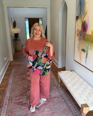 A woman stands in a home hallway holding up a colorful floral tote bag. She is wearing a rust-orange sweatshirt and pink pants, standing on a patterned pink rug next to a white woven bench and a large abstract painting.