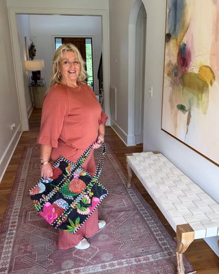 A woman stands in a hallway holding a large, colorful patchwork tote bag. She is wearing a dusty rose top and matching pants.