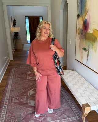 A woman stands in a hallway wearing a dusty rose pink two-piece lounge set consisting of a short-sleeve top and matching wide-leg pants. She is holding a patterned shoulder bag and standing on a large patterned area rug next to a white woven bench and a large abstract painting.