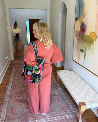 A woman stands in a hallway wearing a matching pink two-piece outfit with wide-leg pants and puff sleeves, carrying a colorful floral tote bag. The setting includes a patterned rug, a bench, and a large abstract painting on the wall.