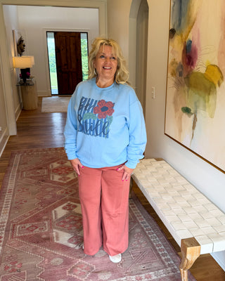 A woman stands in a home hallway wearing a light blue sweatshirt with a red graphic and the words 'RIP UP CARNATION' paired with dusty pink wide-leg pants. She is positioned next to a woven bench and a large abstract painting, with a patterned rug on the floor.