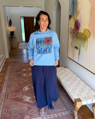 A woman stands in a home hallway wearing a light blue crewneck sweatshirt with a colorful graphic that reads 'HONOR SCHOOL' and a large red flower motif, paired with dark navy wide-leg pants.