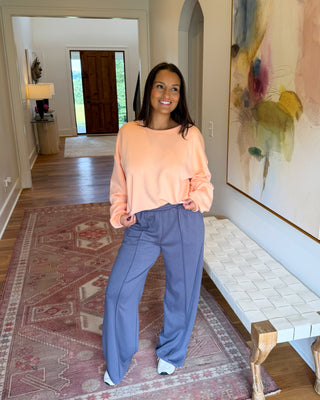 A woman stands in a home hallway wearing a peach long-sleeve top and wide-leg blue pants. The setting features a patterned rug, a woven bench, and a large abstract painting on the wall.