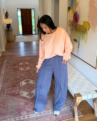 A woman stands in a home hallway wearing a peach crewneck sweatshirt and wide-leg slate blue pants. The setting features a patterned rug, a woven bench, and a large abstract painting on the wall.
