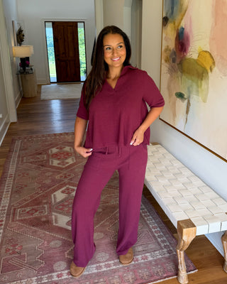 A woman modeling a matching two-piece scrub set in a deep berry purple, standing in a home hallway with hardwood floors, a patterned rug, and a large abstract painting on the wall.