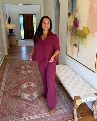 A woman modeling a matching burgundy set consisting of a short-sleeve collared top and matching pants, standing in a home hallway with a patterned rug, a bench, and a large abstract painting.