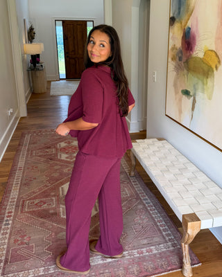 A woman modeling a matching mauve-purple lounge set consisting of a short-sleeve top and wide-leg pants, standing in a home hallway next to a white bench and a large abstract painting.