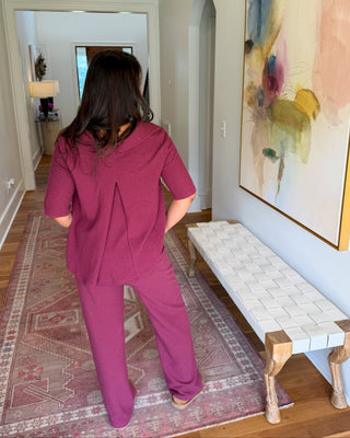 A woman viewed from behind wearing a matching magenta-purple scrub-style top and pants set in a home hallway with a patterned rug, bench, and abstract wall art.
