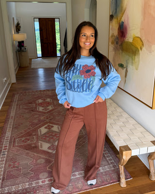 A woman stands in a home hallway wearing a light blue crewneck sweatshirt with a colorful graphic and brown wide-leg pants.
