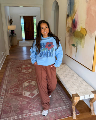 A woman stands in a home hallway wearing a light blue sweatshirt with a colorful graphic and brown pants, posing next to a woven bench and a large abstract painting.