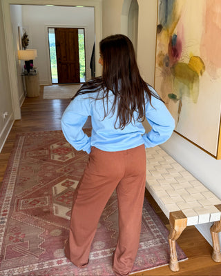 A woman stands in a hallway wearing a light blue long-sleeve top and rust-brown wide-leg pants, viewed from behind. The setting includes a patterned rug, a bench, and a large abstract painting on the wall.