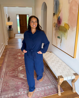 A woman wearing a navy blue long-sleeve jumpsuit with a tie waist stands in a home hallway with hardwood floors, a patterned rug, and a large abstract painting on the wall.